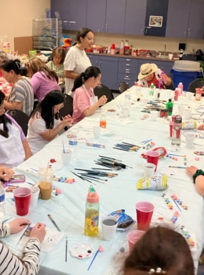 Members and families seated around a long table covered with paint, brushes and craft supplies.