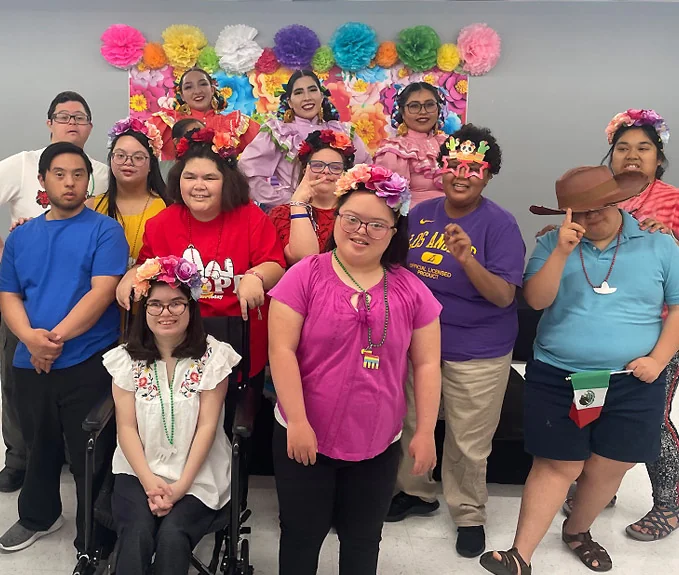 A dozen members posing together in floral crowns in front of a wall of bright paper flowers at a fiesta party.