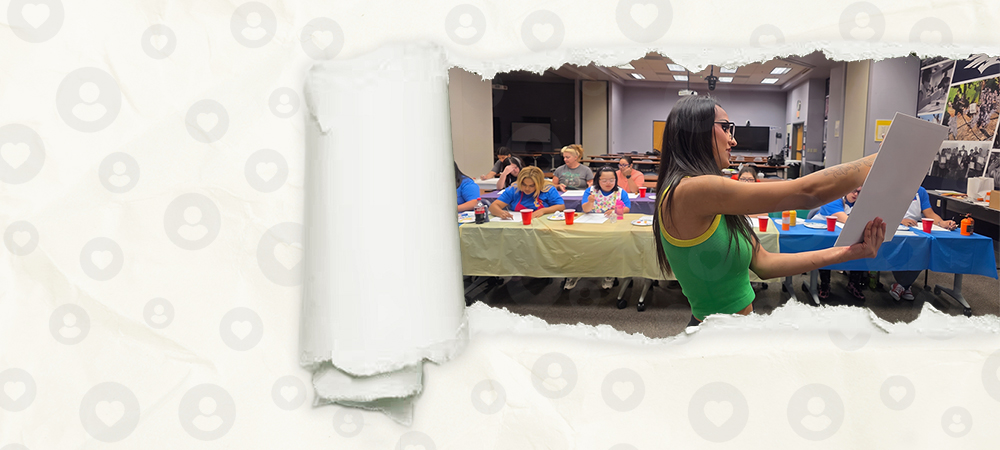 A volunteer holding up a sheet of paper at the front of an arts and crafts class, with members working at tables behind her, seen through a tear in a sheet of paper.